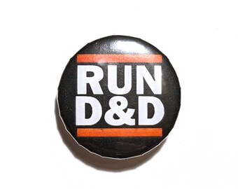 May include: A round, black button with the words "RUN D&D" in bold, white capital letters. The text is separated by a horizontal orange stripe. The button is a fun accessory for fans of tabletop role-playing games.
