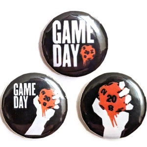 May include: Three black and white button badges with the text "GAME DAY" and a red 20-sided die. The die is held in a white hand on two of the badges.