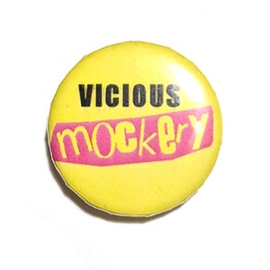 Puede incluir: Un botón amarillo con las palabras "VICIOUS" en negro sobre "mockery" en rosa. El texto está sobre un fondo amarillo. El botón es redondo y brillante.