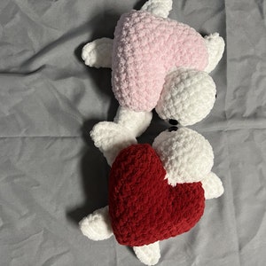 Peut inclure: Deux peluches en forme de cœur crochetées, l'une rose et l'autre rouge, avec des visages blancs en crochet et des yeux noirs.