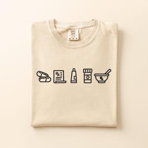 Könnte beinhalten: Ein beiges Kurzarm-T-Shirt mit einer schwarzen Strichgrafik von Apothekenartikeln, darunter zwei Kapseln, ein Rezeptblock, eine Salbentube, eine Medikamentenflasche mit Kreuz sowie ein Mörser mit Stößel und einem Rx-Symbol.