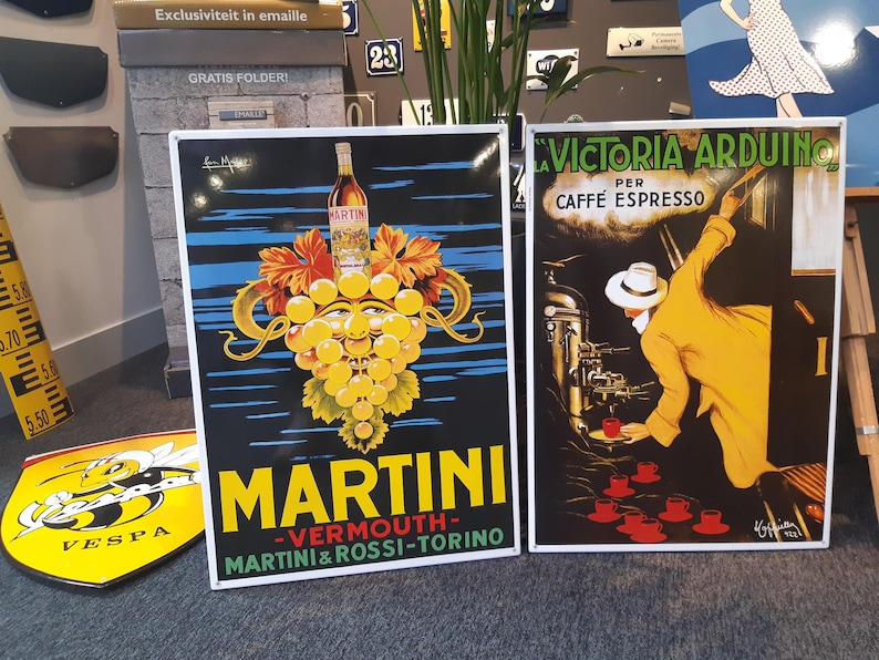 K&ouml;nnte beinhalten: Zwei Vintage-Emaille-Schilder. Das linke Schild ist eine Martini-Werbung mit einer gr&uuml;nen, gelben und wei&szlig;en Trauben-Traube mit einem Gesicht. Das rechte Schild ist eine Victoria Arduino-Kaffeew Werbung mit einem Mann in einem gelben Anzug, der Kaffee gie&szlig;t.
