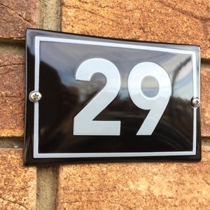 以下が含まれることがあります： 黒い長方形の家の番号プレートで、数字の「29」が銀色で表示されています。 プレートには白い縁があり、2本の銀色のネジでレンガの壁に取り付けられています。