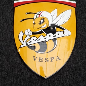 K&ouml;nnte beinhalten: Ein gelbes und schwarzes Vespa-Logo mit einer Cartoon-Biene, die einen Helm tr&auml;gt und einen Vespa-Roller h&auml;lt. Das Logo befindet sich auf einem schildf&ouml;rmigen Hintergrund mit einem rot-wei&szlig;-gr&uuml;nen Rand.