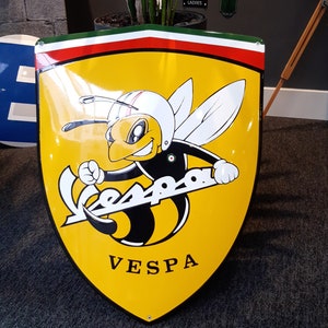 K&ouml;nnte beinhalten: Ein gelbes Metallschild mit einem Vespa-Logo, das eine Cartoon-Biene zeigt, die einen Helm tr&auml;gt. Die Biene fliegt und h&auml;lt das Vespa-Logo. Das Schild hat einen roten, wei&szlig;en und gr&uuml;nen Rand.