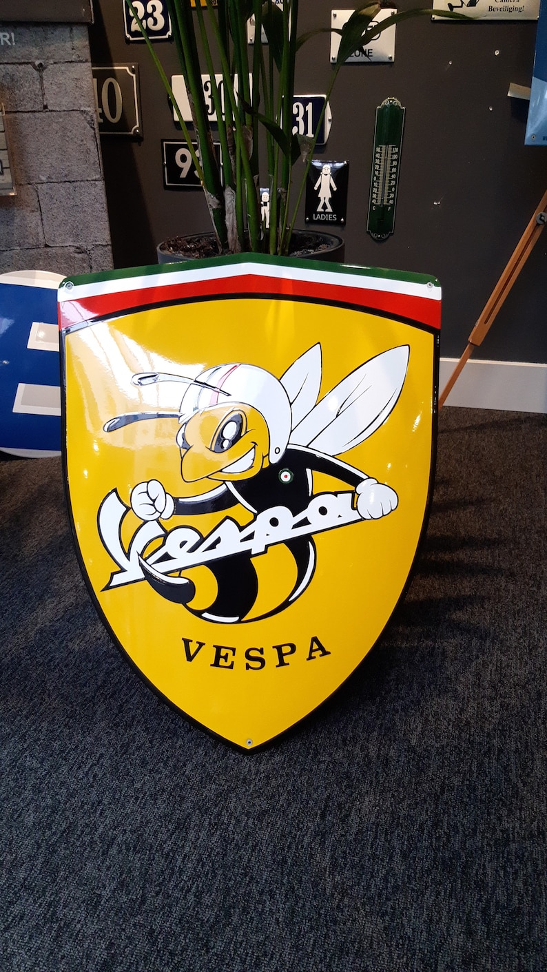 K&ouml;nnte beinhalten: Ein gelbes Metallschild mit einem Vespa-Logo, das eine Cartoon-Biene zeigt, die einen Helm tr&auml;gt. Die Biene fliegt und h&auml;lt das Vespa-Logo. Das Schild hat einen roten, wei&szlig;en und gr&uuml;nen Rand.