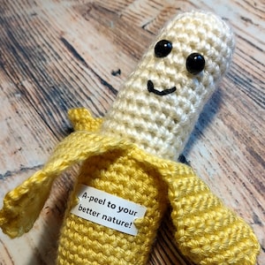 May include: A crocheted banana with a smiling face and black eyes. The banana is yellow and has a brown stem. A white tag is attached to the banana with the text "A-peel to your better nature!"