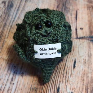 May include: A green crocheted artichoke plush toy with a smiling face and black eyes. The toy is holding a white tag that says "Okie Dokie Artichoke".