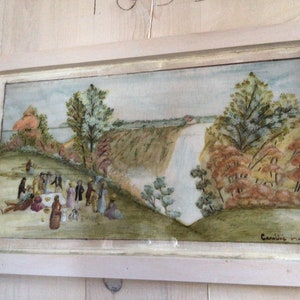 Puede incluir: Una pintura al agua enmarcada que representa una escena de picnic con personas reunidas alrededor de una mesa en un campo de hierba. En el fondo, una cascada cae por un acantilado con &aacute;rboles y vegetaci&oacute;n que la rodean. La pintura est&aacute; firmada "Caroline 191..." en la esquina inferior derecha.