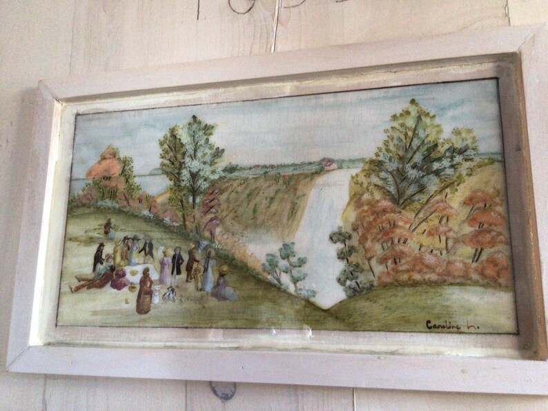 Puede incluir: Una pintura al agua enmarcada que representa una escena de picnic con personas reunidas alrededor de una mesa en un campo de hierba. En el fondo, una cascada cae por un acantilado con &aacute;rboles y vegetaci&oacute;n que la rodean. La pintura est&aacute; firmada "Caroline 191..." en la esquina inferior derecha.