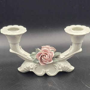 Puede incluir: Un candelabro de cerámica blanca con dos portavelas. Una rosa rosa con hojas verdes está centrada entre los portavelas. El candelabro tiene una base decorativa y un diseño curvo.