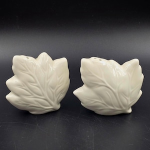 Puede incluir: Dos saleros y pimenteros de cerámica blanquecina. Cada salero y pimentero tiene forma de hoja con vetas detalladas. Los saleros y pimenteros están sobre un fondo oscuro. Artículos de cocina decorativos.
