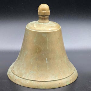 May include: A vintage brass bell with a rounded base and a small handle on top. The bell has a slightly aged appearance with some green patina. The bell is likely made of solid brass and is a classic decorative item.