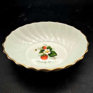 Puede incluir: Un cuenco ovalado de cerámica blanquecina con borde dorado. El cuenco presenta un diseño de fresa en el centro, con las palabras "Strawberry Ripe" debajo. El cuenco tiene un diseño acanalado y probablemente sea para servir.