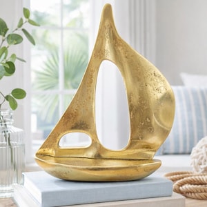 Puede incluir: Una escultura de velero dorada con un gran diseño de vela abierta. La escultura se encuentra sobre una pila de libros. Las palabras "Styling Inspiration" están en la parte inferior de la imagen. Un jarrón de vidrio transparente con vegetación está a la izquierda.