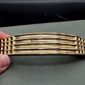 Może przedstawiać: Złoty uchwyt do szafki o zakrzywionym kształcie. Uchwyt posiada szereg poziomych grzbietów i kwadratowych wzorów kratowych na każdym końcu. Okucia są prawdopodobnie wykonane z metalu i mają wygląd vintage.