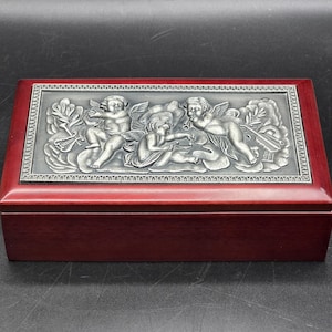 Könnte beinhalten: Eine rechteckige Schmuckschatulle aus Holz mit dunkelroter Oberfläche. Der Deckel zeigt ein silberfarbenes, geprägtes Design von Engeln mit Musikinstrumenten und floralen Akzenten. Die Box ist geschlossen und steht auf einer dunklen Oberfläche.