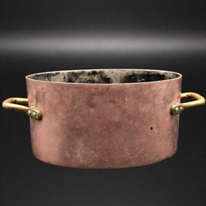 May include: A copper cooking pot with brass handles. The pot has a rounded shape and a slightly flared rim. The exterior is a warm, reddish-brown color, while the interior shows signs of use. The handles are attached with rivets.