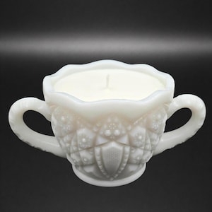 Puede incluir: Una vela blanca en una taza de té decorativa de estilo vintage con dos asas. La vela tiene una superficie lisa y blanca y una sola mecha. La taza de té tiene un exterior texturizado y estampado.