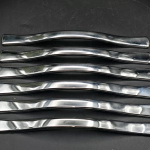 May include: Six polished, silver-toned cabinet handles with a gently curved, ergonomic design. The handles have a smooth, reflective surface, and are arranged in parallel rows against a dark background. These are suitable for kitchen or bathroom cabinets.