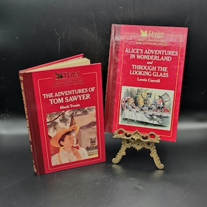 Può includere: Due libri vintage con copertina rigida, "Le avventure di Tom Sawyer" di Mark Twain e "Alice nel paese delle meraviglie" di Lewis Carroll, sono esposti. I libri hanno copertine rosse con scritte e illustrazioni dorate. "Alice" è su un piccolo e ornato supporto dorato.