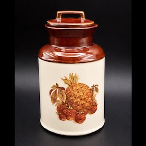 May include: A vintage ceramic jar with a lid, in brown and cream. The jar is decorated with a pineapple and strawberry fruit design. The lid has a handle. This decorative jar is suitable for kitchen storage. The jar measures approximately 20 cm in height.