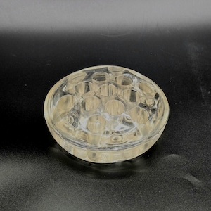 May include: Clear glass flower frog with multiple holes. The round frog is designed to hold flower stems in place. The glass has a slight yellow tint and is sitting on a dark surface.