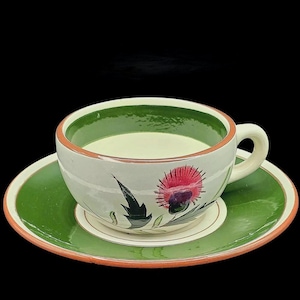 Peut inclure: Un ensemble tasse et soucoupe en céramique orné d'un chardon rose et violet peint à la main sur l'extérieur. La tasse présente une base crème avec un bord intérieur vert foncé et un liseré couleur terre cuite sur la tasse et la soucoupe.