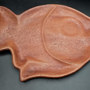 Puede incluir: Una bandeja de madera con forma de pez. La bandeja es de madera marrón rojiza con vetas visibles. La bandeja con forma de pez tiene un diseño curvo y es adecuada para servir alimentos.