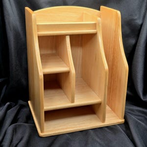 May include: A light-colored wooden desk organizer with multiple compartments and shelves. The organizer has a vertical design with a curved top and sides, and is made of natural wood.