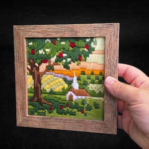May include: A square needlepoint embroidery in a light brown wooden frame depicting a landscape with a large tree featuring red fruit, a small white building with a brown roof, yellow fields, and a soft orange and cream sky in the background.