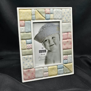May include: A white 4x6 photo frame with a patchwork design in pastel colors. The frame holds a black and white photo of a baby wearing a checkered hat. The frame is made by Burnes of Boston.