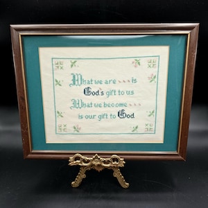 May include: Framed cross-stitch artwork with a brown wooden frame, teal matting, and a gold-coloured stand. The cross-stitch features the text "What we are is God's gift to us. What we become is our gift to God" in black script.