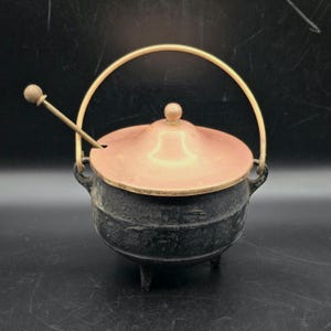 Puede incluir: Un caldero de hierro fundido negro con tapa y asa de color cobre. Una pequeña cuchara de madera descansa sobre la tapa. El caldero tiene tres patas pequeñas y un asa de latón. Un artículo de cocina decorativo.