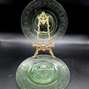 Può includere: Due piatti di vetro verde con motivi floreali sono esposti su un supporto decorativo color oro. I piatti hanno un design centrale e un bordo dettagliato. Il supporto è ornato, con un design dettagliato. Lo sfondo è nero.