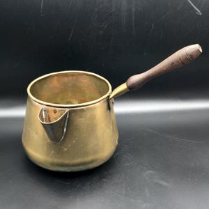 Puede incluir: Una cafetera de latón vintage con un mango de madera. La cafetera tiene una base redondeada, un pico y un mango largo. El latón tiene un color dorado cálido y el mango de madera es de color marrón oscuro.