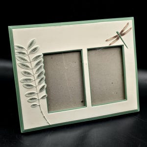 May include: A double photo frame with a cream-colored center and a light green border. The frame is decorated with a raised leaf design on the left and a dragonfly on the upper right. The frame is designed to hold two photos.
