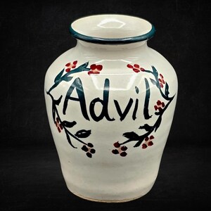 May include: A cream-colored ceramic jar with a dark teal rim. The word Advil is hand-painted in large dark teal letters on the front, surrounded by a decorative painted vine motif featuring dark teal leaves and small red berries.