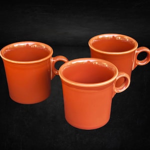 Puede incluir: Tres tazas de cerámica de color naranja sólido con un acabado liso y brillante. Cada taza presenta un cuerpo cilíndrico, un asa redondeada en el lateral y un sutil detalle de anillo horizontal cerca del borde superior. Las tazas están agrupadas.