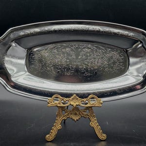 Puede incluir: Una bandeja ovalada plateada con grabados florales ornamentados. La bandeja está sostenida por un soporte decorativo de metal dorado. Los bordes de la bandeja tienen un diseño festoneado.
