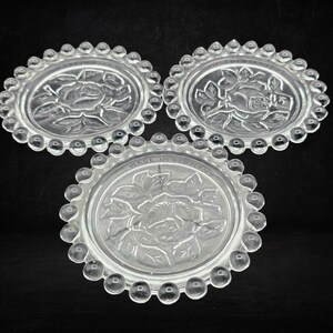 Könnte beinhalten: Drei kreisförmige Untersetzer aus klarem Glas mit einem erhabenen Rosenmuster in der Mitte und einem Perlenrand. Jeder Untersetzer hat ein strukturiertes Blumendesign und der Text MADE IN GERMANY ist auf der Oberfläche des unteren Untersetzers eingeprägt.