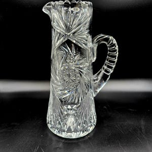Op de afbeelding: Een heldere, geslepen glazen kan met een decoratief patroon van sterren. De kan heeft een lange, slanke body, een tuit en een geribbelde handgreep. Het glas is ingewikkeld gesneden met geometrische ontwerpen, waardoor een sprankelend effect ontstaat.