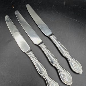 May include: Three stainless steel butter knives with decorative floral handles. The blades are smooth, and the handles feature an embossed pattern. The knives are displayed on a dark surface.