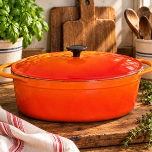 May include: An oval enameled cast iron Dutch oven in a vibrant gradient orange color. The pot features two side handles and a matching lid with a black knob. The exterior transitions from a deep red-orange at the rim to a bright orange base.