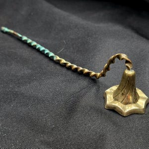 May include: A vintage brass candle snuffer with a spiral handle and a bell-shaped head. The handle transitions from a green-blue hue to a golden brass color. The bell-shaped head has a star-like base.