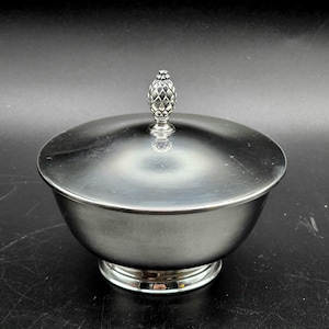 Könnte beinhalten: Eine silberfarbene Schüssel mit Deckel. Der Deckel ist mit einem dekorativen Zierrat in Form einer Ananas versehen. Die Schüssel hat einen runden Boden und eine polierte, reflektierende Oberfläche. Die Schüssel ist wahrscheinlich aus Metall.