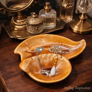 May include: A leaf-shaped ceramic trinket dish with a glossy, variegated amber and cream glaze. The dish contains two necklaces, one with a silver-toned pendant and another featuring a long chain with a smooth, white teardrop-shaped stone pendant.