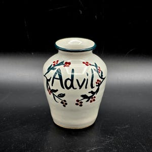 May include: A white ceramic vase with a dark teal rim and the word "Advil" in black script. The vase is decorated with green leaves and red berries.