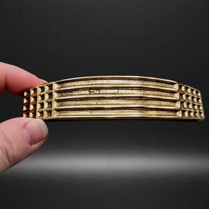 May include: A gold-coloured, rectangular metal object with horizontal ridges and square-patterned ends. The object is held in a hand against a dark background. The design is reminiscent of Art Deco.