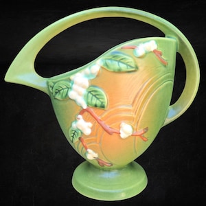 May include: A ceramic pitcher featuring a green and orange gradient finish with a raised relief design of white flowers and green leaves on brown branches. The vessel has a wide, arched handle and a circular pedestal base, set against a black background.